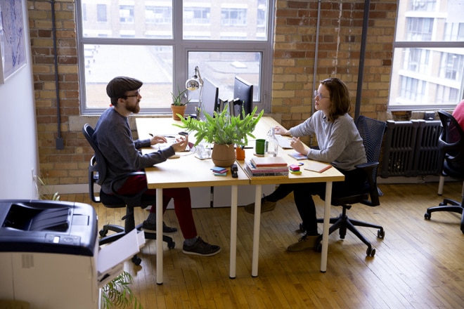 Jay and Kaila sit at their workspaces in a bright design studio with brick walls and hardwood floors, speaking to each other.
